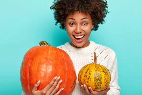 pleased smiling curly woman chooses pumpkin for halloween holds big and small squash wears white sweater - Маленькие хитрости приготовления пищи