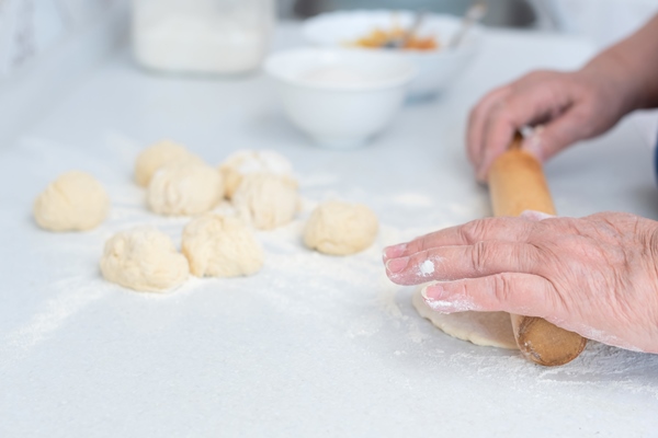 senior woman hands rolling out the dough with a rolling pin on a white kitchen table with blurred grated apple and sugar on background selective focus process of making pies with apple filling - Шанежки на кефире с картошкой