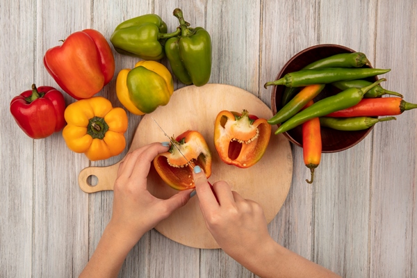 top view of female hands cutting orange bell peppers on a wooden kitchen board with knife on a grey wooden surface - Аджика с яблоками
