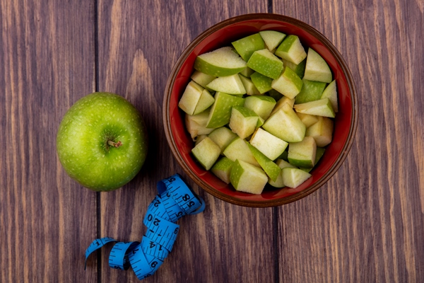 top view of whole apple with chopped apple slices on a red bowl on a wooden surface - Пирог с яблоками