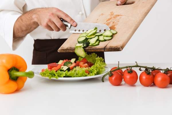 young cook man in uniform cutting vegetable salad on wooden board isolated over white wall - Овощи, бобовые, грибы: полезные советы