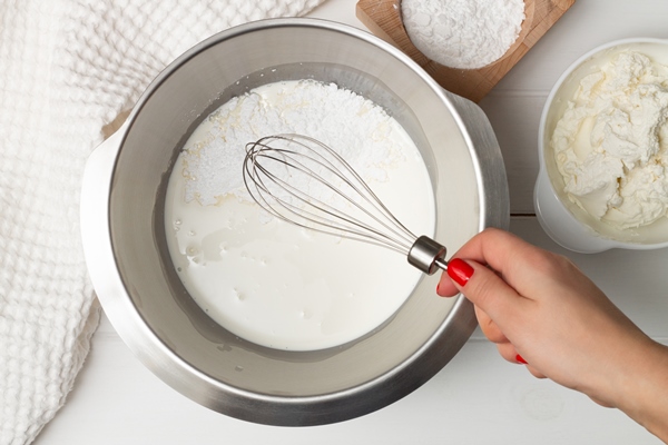 a woman s hand with a whisk over a bowl of whipping cream and powdered sugar near a towel and bowls of powdered sugar and ricotta flat lay - Пасха старинная