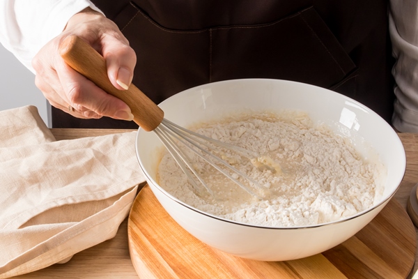 a womans hand stirs the flour with a whisk in a bowl with the ingredients for the dough butter eggs milk a can of flour on the kitchen table - Постные блинчики с капустой и баклажанами