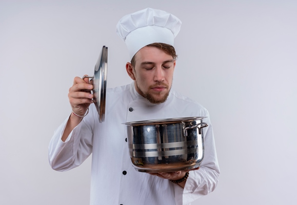 a young bearded chef man wearing white cooker uniform and hat smelling a cooking pan on a white wall - Пасха варёная