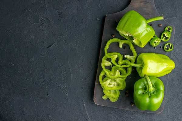above view of whole cut chopped green peppers on dark color wooden cutting board on the left side on black surface - Салат из перца и баклажанов