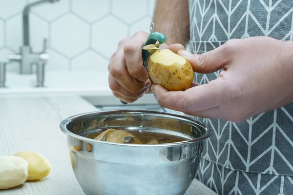 cropped photo of man wearing grey apron peeling potato with green peeler over metal bowl organic waste management - Суп с фрикадельками из индейки