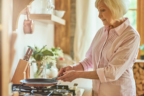 elderly lady holding a frying pan on the stove in the kitchen - Постные закусочные блинцы на соевом молоке