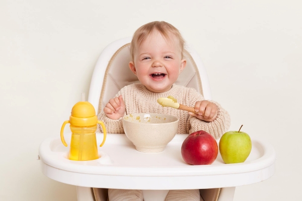 image of deligted laughing infant baby girl dresses in beige jumper sitting in high chair and eating isolated over white background holding spoon in hands enjoying tasty fruit puree - Особенности диеты при аллергических заболеваниях