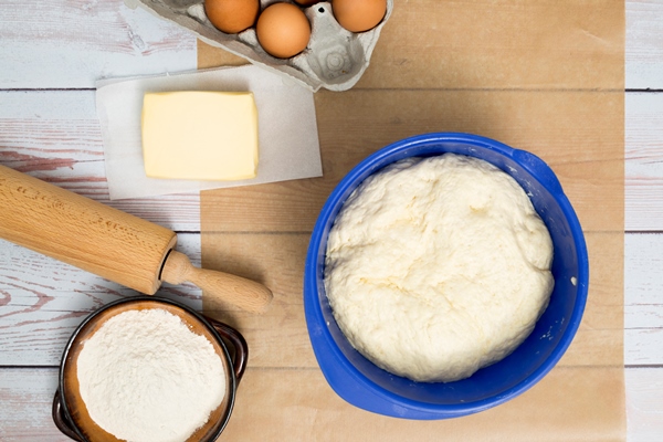 kneaded dough in blue bowl eggs butter flour and rolling pin on wooden desk - Пасхальный хлеб по-голландски