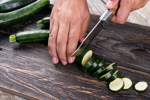 man cutting fresh zucchinis into slices on a cutting board on a wooden table - Запеканка из кабачков и цветной капусты