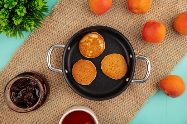 top view of pancakes in pan and strawberry jam tea apricots on sackcloth on blue background - Постные овсяные блины