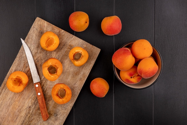 top view of pattern of half cut apricots with knife on cutting board and whole ones in bowl and on black background - Абрикосовый компот с апельсинами