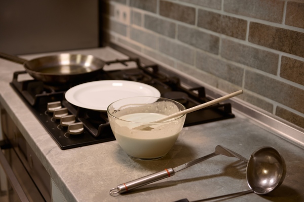 wooden spoon in glass bowl with pancake batter stainless steel kitchen utensils on kitchen countertop white plate and frying pan on a black stove - Постные ванильные блинцы