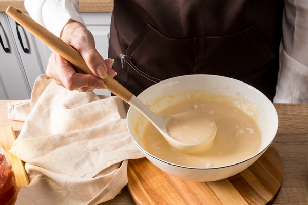 a faceless woman holds a ladle of cooked liquid pancake dough over a bowl kitchen countertop with ingredients for further cooking - Оладьи с корицей и лимонной цедрой