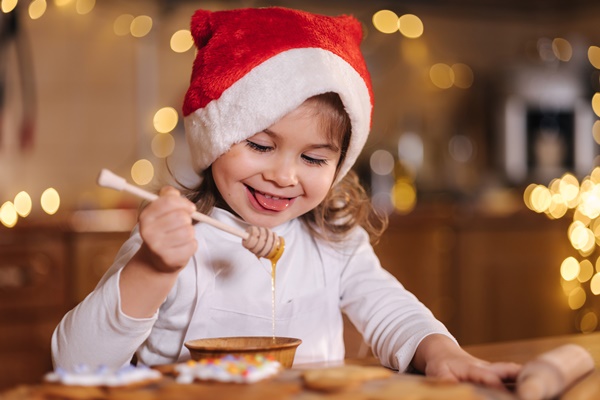 little girl in red santa hat eating honey using honey stick kitchen decorated by fairy lights - Медовые пряники
