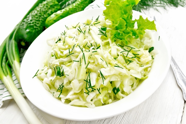 salad of fresh cabbage green onions and cucumber with vinegar and vegetable oil dressing in a plate towel dill and fork on light wooden board background - Свежая шинкованная капуста