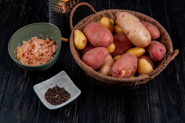 side view of whole potatoes in basket and grated ones in bowl with black pepper seeds and grater on wooden table 1 - Клёцки с грибами