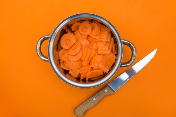 sliced carrots in a metal saucepan with a knife on an orange background - Морковь тушёная постная