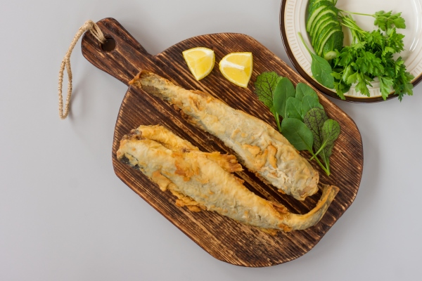 top view of fried fish navaga served on wooden cutting board with lemon vegetables and greens on gray background - Навага жареная, постный стол