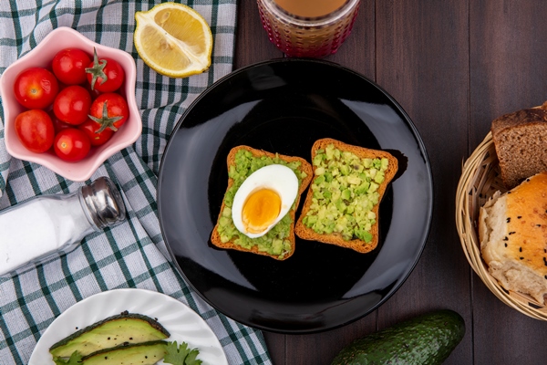 top view of toast bread slices with avocado pulps and egg on a black plate with tomatoes lemon on checked tablecloth and wooden surface - Перекус "Отличник"