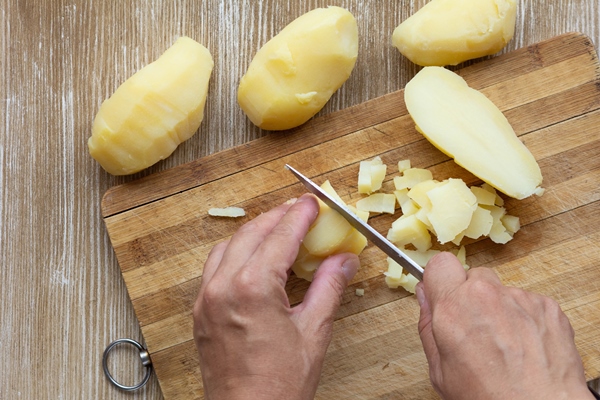 top view of woman hands cutting scrubbed boiled potatoes in jackets using knife on the wooden background - Картофельный салат с маринованными огурцами