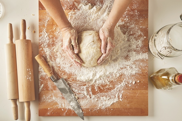 top view on white table with isolated wooden board with knife two rolling pins bottle olive oil transparent jar with flour woman hands hold prepared dough for pasta or dumplings - Ватрушки с вареньем или яблоками