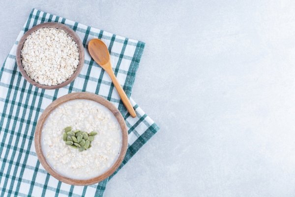 delicious breakfast serving of pepitas topped oatmeal on folded tablecloth next to a bowl of oat and a wooden spoon on marble background - Суп овсяный с яблоками