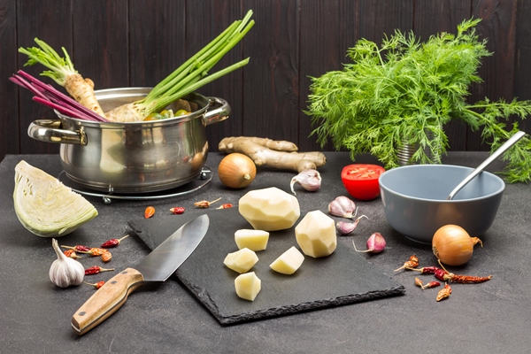 peeled potatoes and knife on cutting board parsley roots and beets in saucepan greens and vegetables on table black background - Суп-рассольник с перловкой