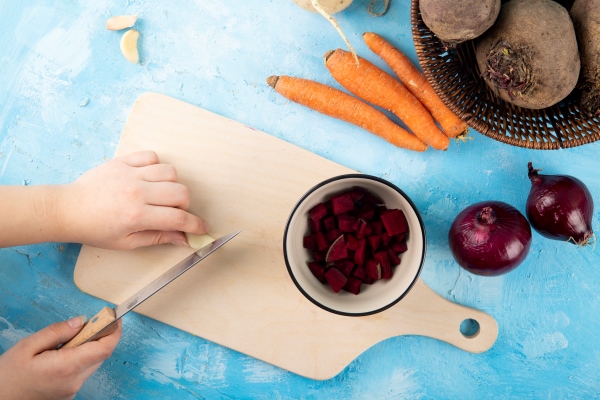 top view woman cuts garlic on a board with chopped beets in a cup with carrots and red onions on a table - Борщ украинский