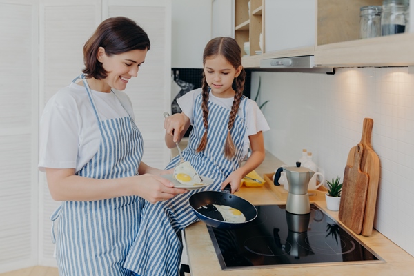 photo of cheerful busy mum and her daughter pose near stove serve breakfast for family fry eggs on frying pan wear aprons pose in domestic kitchen - Готовим яичницу вместе с детьми