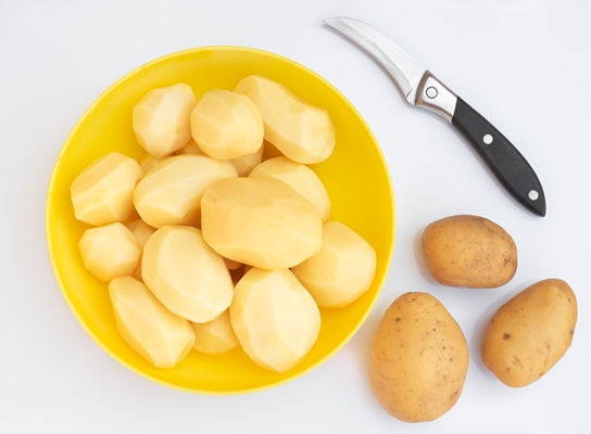 peeled potatoes in a yellow cup on a light surface nearby there is a knife and potato tubers cooking - Драники с яйцом