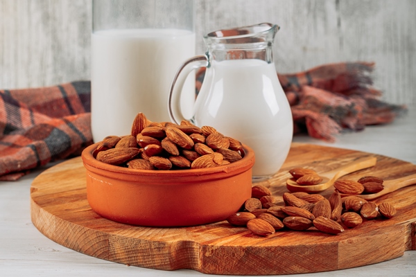 side view milk carafe and bottle with bowl of almonds on wooden board on white wooden background horizontal - Пряный миндальный горячий шоколад