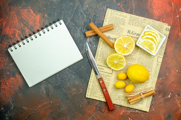 top view fresh lemons cumcuats cinnamons lemon slices in small bowl knife on newspaper notebook on dark red table - Безалкогольный глинтвейн на чае и соке