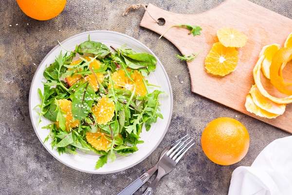 diet vegetarian vitamin salad of orange slices and a mix of arugula chard and mizun leaves on a plate and a cutting board with peeled orange on the table top and vertical view - Винегрет из фруктов и овощей