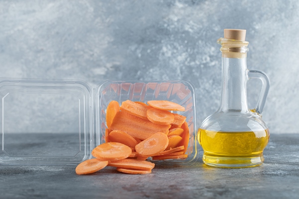pile of sliced carrots in plastic container and bottle of oil over grey background - Творожники с морковью