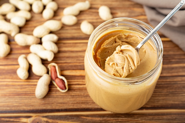 creamy peanut paste in open glass jar peanut butter in spoon peanuts in the peel scattered on the brown wooden table - Применение арахисовой пасты