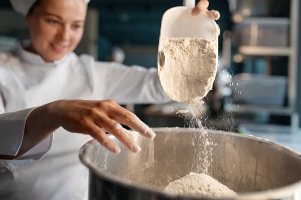 professional female confectioner wearing white uniform putting flour into big metal bowl standing near table in confectionery closeup view selective focus - Монастырская кухня: галушки по-охотничьи, лимонное печенье