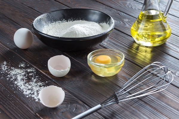 sieve on black bowl with flour glass bottle with oil eggs eggshell broken egg in plate metal whisk wooden spoon with flour - Блинцы на йогурте