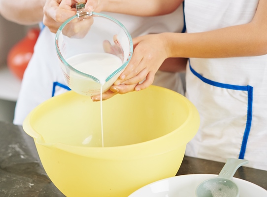 smiling mother and daughter pouring jar of warm milk in big plastick bowl when making pancake dough - Панкейки с корицей