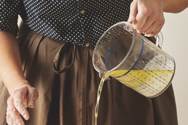 woman pours water with olive oil from measure cup to flour on board to prepare dough for pasta or dumplings cooking guide presentation - Монастырская кухня: вареники с яблоком и изюмом, морковное печенье