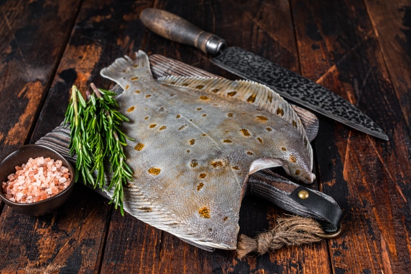 raw plaice flatfish fish on butcher board with knife dark wooden background top view - Камбала с помидорами