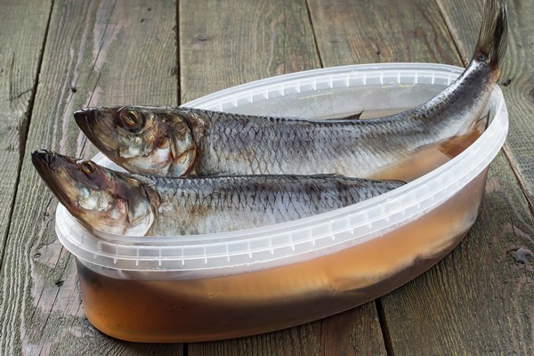 two salted herrings in plastic container with brine and spices on a wooden table - Консервирование пищевых продуктов