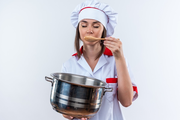 with closed eyes young female cook wearing chef uniform holding saucepan trying soup with spoon isolated on white background - Питание в походе