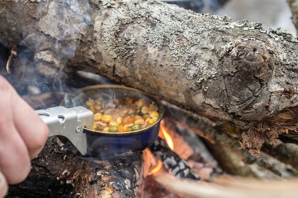 man prepares a meal over a campfire while holding a metal can over the fire - Мясо с овощами по-походному