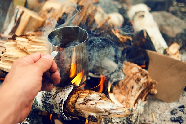 man traveler hands holding cup of tea near the fire outdoors hiker drinking tea from mug at camp coffee cooked over a campfire on the nature adventure travel tourism and camping concept - Кофе "Походный"