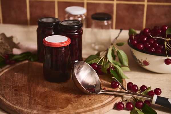 freshly made jam poured into jars stand next to ladle fresh green cherry berries with green leaves on wooden board in rustic cozy home wooden kitchen - Вишнёвое варенье