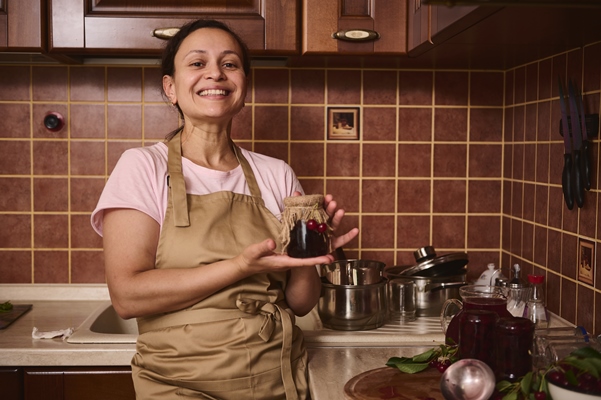 pleasant woman housewife wearing chef apron smiles looking at camera holding a glass jar with freshly made cherry jam - Вишнёвое варенье