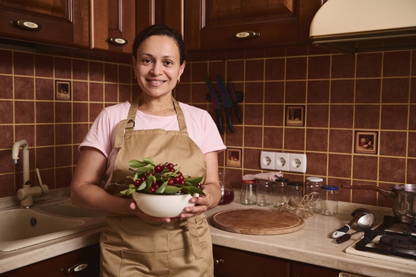 woman holds a bowl full of freshly picked cherry berries looks at camera standing at home wooden kitchen background - Вишнёвое варенье