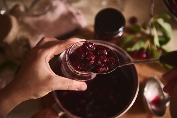 hands of a housewife holding a sterilized glass jar and filling it with homemade freshly brewed cherry berry jam over a saucepan in a home kitchen - Вишнёвое варенье с косточками
