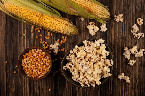 top view of a bowl of popcorns with corn kernels on a wooden bowl with fresh corns isolated on a wooden wall - Воздушная кукуруза (попкорн)